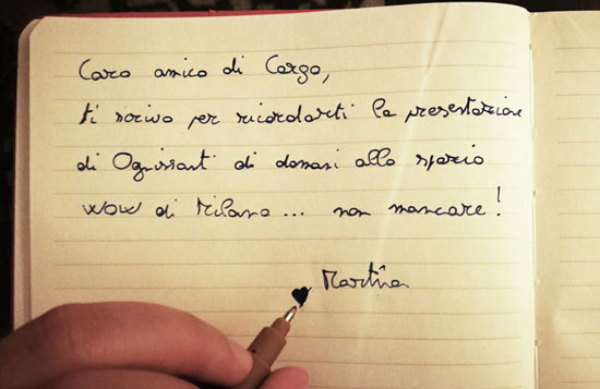Una mano che scrive: caro amico di Cargo, ti scrivo per ricordarti la presentazione di Ognissanti di domani allo spazio WOW di Milano, non mancare!